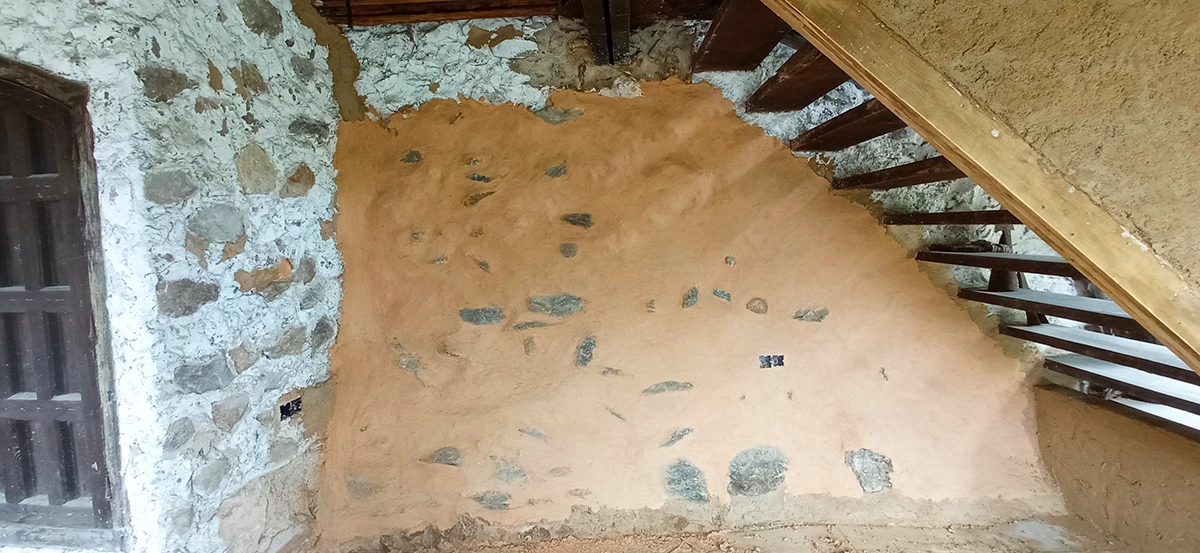 Muro de mampostería tradicional restaurado con revestimiento de tierra bajo una escalera de madera en una vivienda de la Sierra de Aracena.